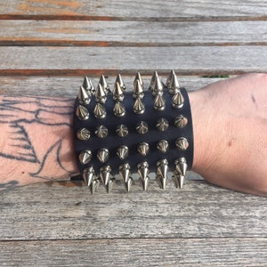 May include: A black leather wrist cuff with silver pyramid-shaped studs. The cuff is worn on a person's wrist.