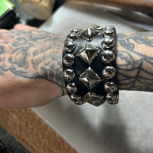Op de afbeelding: Zwarte leren armband met zilverkleurige piramide- en ronde studs. De brede armband omsluit de pols. De studs zijn gelijkmatig verdeeld en geven het accessoire een stoere, edgy uitstraling.
