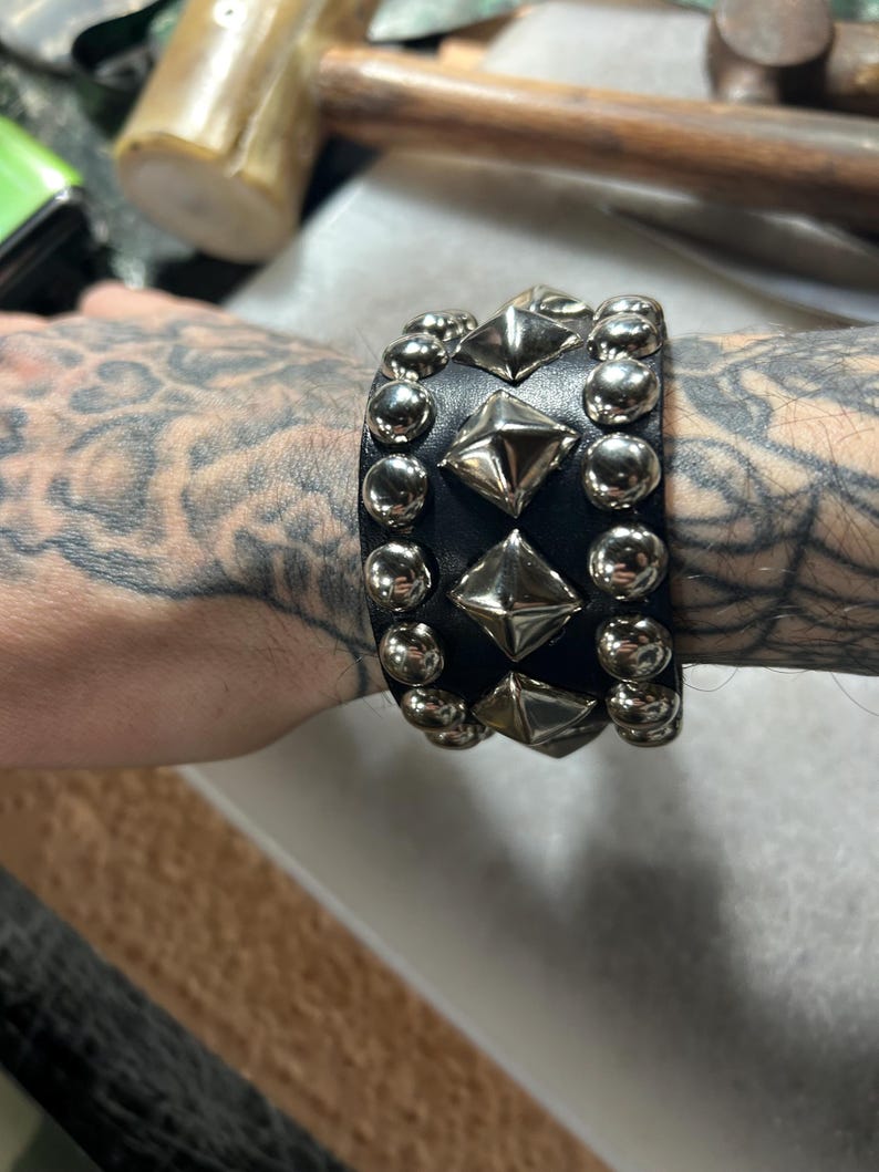 Op de afbeelding: Zwarte leren armband met zilverkleurige piramide- en ronde studs. De brede armband omsluit de pols. De studs zijn gelijkmatig verdeeld en geven het accessoire een stoere, edgy uitstraling.