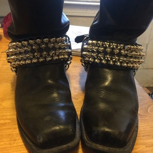 Puede incluir: Un par de botas de cuero negro con correas de tobillo tachonadas de plata. Las correas están conectadas por una cadena de plata.