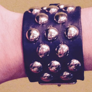 Könnte beinhalten: Ein schwarzes Lederarmband mit silbernen Nieten. Das Armband hat drei Reihen Nieten, jede Reihe mit fünf Nieten.