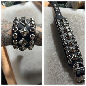 Op de afbeelding: Zwarte leren armband en halsband versierd met zilverkleurige piramide- en ronde studs. De armband heeft drie rijen studs, de halsband twee. De armband wordt getoond op de arm van een persoon.