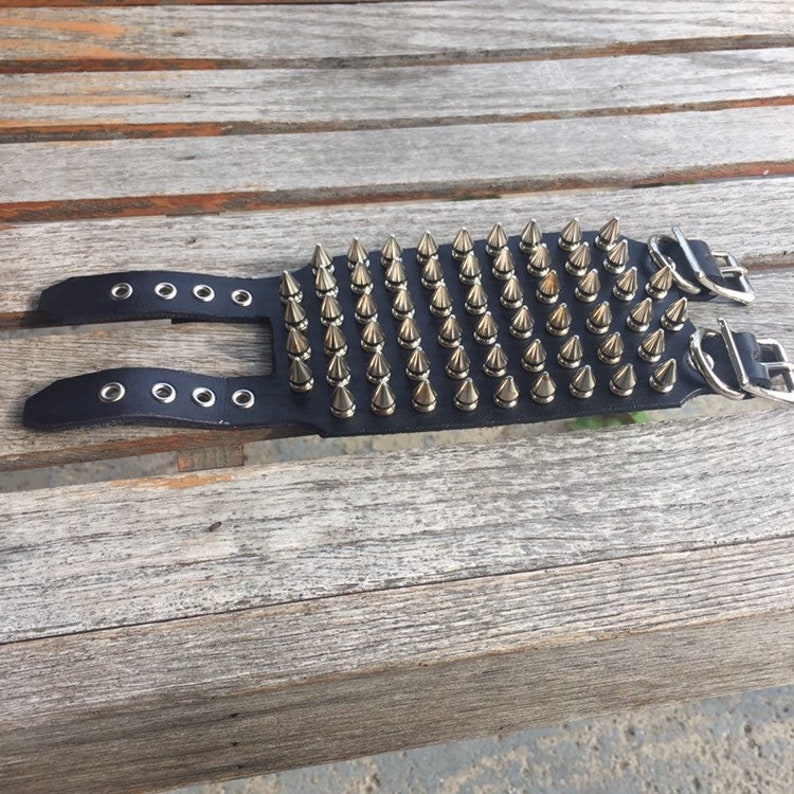 May include: A black leather bracelet with silver pyramid-shaped studs. The bracelet has two adjustable straps with metal buckles.