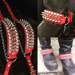 Puede incluir: Dos correas de cuero rojo con tachuelas y púas plateadas. Las correas tienen un detalle de cadena y están diseñadas para usarse alrededor de los tobillos. Las correas se muestran en un par de botas negras.