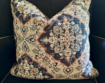 May include: Decorative throw pillow with a detailed, ornate pattern in navy blue, beige, and rust colors. The pillow is square-shaped and appears to be made of a woven fabric, adding texture to the design. The pillow is placed on a dark blue surface.