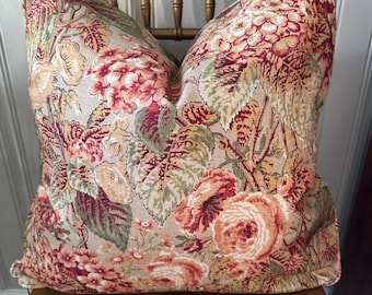May include: A floral pillow with a beige background and a pattern of red, pink, and green flowers. The pillow is sitting on a wooden chair.