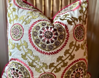 May include: A decorative throw pillow with a white background and intricate embroidery. The design features circular patterns in shades of red, brown, and gold, with a pink border. The pillow is placed on a wooden chair.