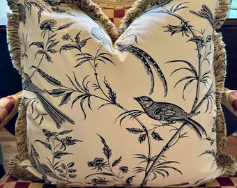 May include: Square decorative pillow with a cream background and a black botanical print featuring birds and foliage. The pillow has a fringed edge in a neutral tone. The pillow is placed on a chair with a red and gold checkered pattern.