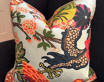 May include: A decorative pillow with a light blue background and a colorful floral and bird design. The pillow features orange, red, yellow, and dark blue accents. The pillow is placed on a dark blue chair.