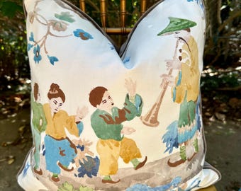 May include: A square decorative throw pillow featuring a white background with a colorful illustration of three figures in traditional attire. One figure plays a long wooden horn while two others dance. The pillow has a dark gray piped edge.