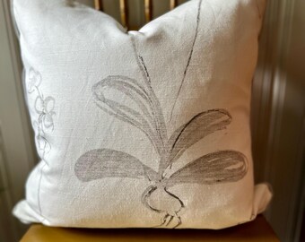 May include: A square, off-white decorative pillow with a botanical design. The pillow features a large, stylized gray plant illustration on the front. The pillow is resting on a wooden chair with a gold finish.