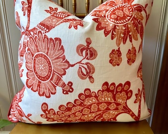 May include: A decorative throw pillow with a white background and a large, repeating floral pattern in shades of red and coral. The pillow is square and has a red trim. The pillow is placed on a wooden chair.