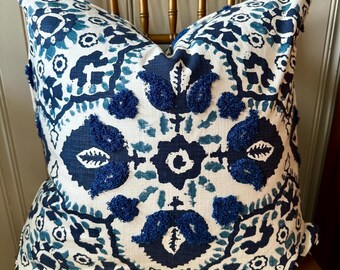 May include: A white throw pillow with a blue and teal patterned design. The pillow has blue pom-poms around the edges and a geometric pattern in the center.