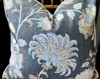 May include: Decorative pillow with a dark gray background and a large floral pattern in shades of blue, beige, and peach. The pillow has a white border and is placed on a gold surface.