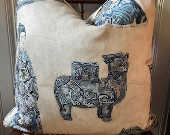May include: A square throw pillow with a cream-colored fabric base featuring a printed design of a person in a blue patterned robe and a stylized four-legged creature with blue and grey markings, including a small rectangular structure on its back.