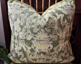 May include: A decorative square pillow with a green and white floral pattern. The pillow has a yellow trim and is placed on a wooden chair. The pillow's design features a detailed, repeating pattern.