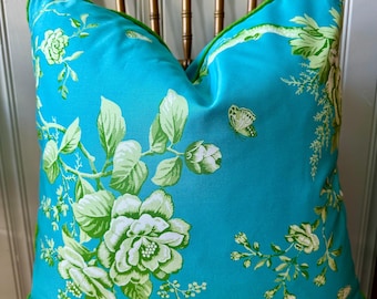 May include: A turquoise blue pillow with a green floral pattern. The pillow has a green border and is sitting on a wooden chair.