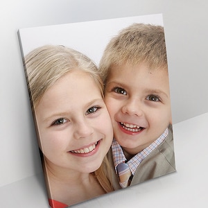 May include: A canvas print featuring a close-up of two smiling children. The girl has long blonde hair and the boy has short blonde hair. The children are wearing casual clothing. The print is displayed at an angle against a white background.