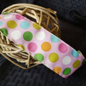 May include: A pink ribbon with colorful polka dots in green, yellow, blue, and pink. The ribbon is wrapped around a woven ball. The back of the ribbon is green.