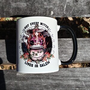 May include: White ceramic mug with a black handle and rim. The mug features a witch illustration with the text "NOT EVERY WITCH LIVES IN SALEM" in a circular design. The witch is depicted with a cauldron and a book.