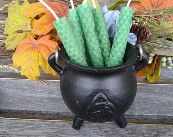 May include: A black cast iron cauldron filled with green honeycomb candles. The cauldron has three legs and two handles. The candles have white wicks and are surrounded by fall foliage.