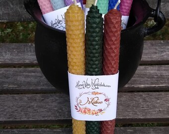 May include: A collection of multi-colored beeswax taper candles with a textured honeycomb pattern. Three candles in the foreground are wrapped in a white paper band labeled HoneyVineMagickals.com and Mabon with an illustration of pumpkins and leaves.