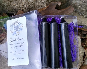 May include: A clear plastic box containing four black candles with white wicks, nestled in purple paper shreds. A white card with blue floral accents and the words "Black Candles" is visible.