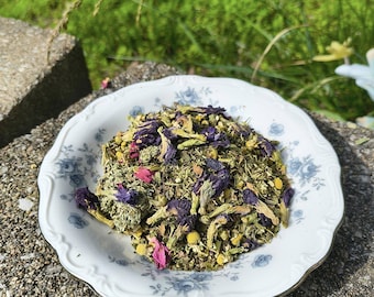 May include: A white, scalloped-edge plate with blue floral designs holds a mix of dried herbs and flowers. The blend includes purple, pink, and yellow blossoms, along with green leaves. The plate sits on a stone surface, with green grass in the background.