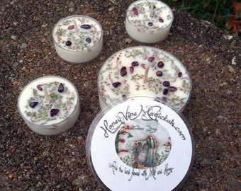 Könnte beinhalten: Eine Ansammlung kleiner, weißer Teelichter, die jeweils mit dunkelroten Steinen und grünen Kräutern verziert sind, wird gezeigt. Eine Kerze trägt ein Etikett mit dem Text "Honey Vine Magickals". Die Kerzen stehen auf einer strukturierten Oberfläche.