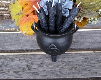 May include: A black cast iron cauldron filled with five black textured candles with white wicks. The cauldron sits on a weathered wooden surface, with a fall-themed wreath in the background, featuring colorful leaves and flowers.