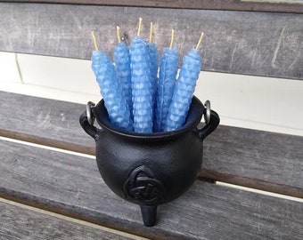 May include: A black cauldron filled with light blue, textured candles with yellow wicks. The cauldron has a Celtic knot design and sits on a wooden bench. The candles are cylindrical and have a honeycomb-like pattern.