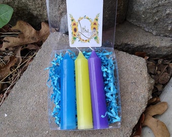 May include: A clear plastic box containing three colorful candles: blue, yellow, and purple. The candles are surrounded by blue shredded paper. A white card with the text "HoneyWineMagickals.com" and "Litha" is visible.
