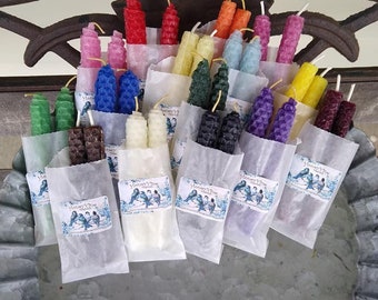 May include: A display of colorful, textured candles in various hues, including red, yellow, blue, and green. The candles are arranged in small paper bags, each with a label featuring a bird design. The candles are displayed in a decorative metal container.
