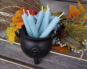May include: A black cauldron with a Celtic knot design holds several light blue hexagonal candles. The candles have a textured surface and white wicks. The cauldron is surrounded by fall foliage, including orange, yellow, and green leaves, and sits on a wooden surface.