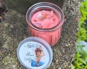 May include: A pink candle in a clear glass jar with a lid. The candle is decorated with crystals and small stones. The lid has a label with the text "Heart Aphrodite" and an illustration of a woman.