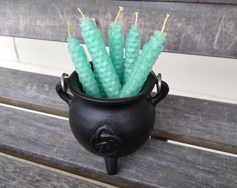 May include: A black cast iron cauldron filled with six light teal honeycomb-patterned candles with yellow wicks. The cauldron has a Celtic knot design and sits on a weathered wooden bench.