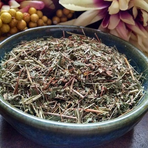 May include: A blue ceramic bowl filled with dried herbs and leaves. The herbs are a mix of green and brown, with some reddish stems. The bowl is set against a blurred background of flowers and a dark wooden surface.