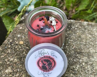 Könnte beinhalten: Eine rote Kerze in einem Glas, verziert mit einem Vogelschädel und anderen Elementen. Der Deckel, neben dem Glas platziert, zeigt den Text "Magpie's Curios" und "Morrighan" in einem kreisförmigen Design.