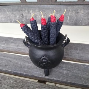 May include: A black cast iron cauldron filled with black candles with red tips. The cauldron has a Celtic knot design on the front and sits on a wooden bench. The candles have a textured, honeycomb-like pattern.
