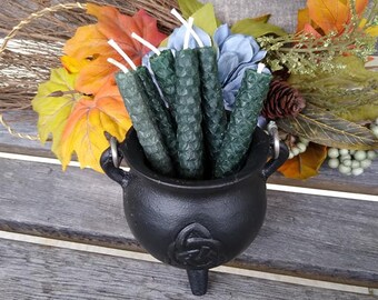 May include: A black cast iron cauldron filled with dark green hexagonal candles. The cauldron has a Celtic knot design and sits on a wooden bench. Autumn leaves and flowers surround the cauldron.