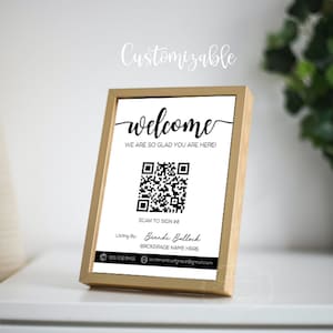 May include: A white sign in a gold frame with the text "welcome" in black cursive font. The sign also includes the text "We are so glad you are here!" and a black and white QR code with the text "Scan to sign in!" below it. The sign also includes the text "Listing By: Brandi Bullock" and "Brokerage Name Here" in black text. The sign also includes the text "910.302.8431" and "sentimentsofgrace@gmail.com" in black text.