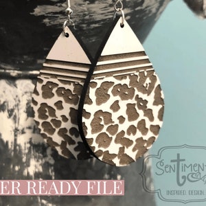 May include: A pair of laser cut wooden earrings with a white and brown leopard print design. The earrings are shaped like teardrops and have a thin black border. The earrings are hanging from silver ear wires.