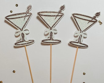 May include: Three martini-shaped cupcake toppers with glittery white interiors and brown outlines. Each topper has a bow and a toothpick stem. A small olive-like detail sits atop each glass. Gold confetti dots the background.