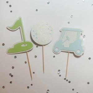May include: Three golf-themed cupcake toppers. One is a green flag on a green base, another is a white golf ball with dimples, and the third is a light blue golf cart. All are attached to wooden sticks.
