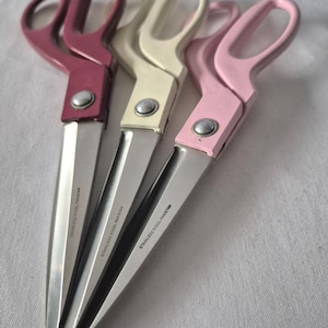 May include: Three pairs of scissors with stainless steel blades and colored handles. The handles are in shades of pink, cream, and burgundy. Each pair has a silver rivet and the text "STAINLESS STEEL PAKISTAN" printed on the blade.