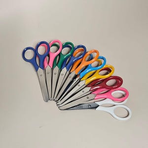 May include: A set of eleven pairs of scissors arranged in a fan shape, each featuring stainless steel blades and plastic handles in various colors including blue, pink, green, orange, yellow, maroon, and white, with the brand name TAMSCO visible.