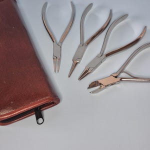 May include: A set of silver metal pliers with various head shapes, including needle-nose and cutting pliers, next to a brown leather case with a zipper. The tools are arranged on a light blue surface.