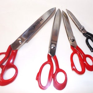 May include: Four pairs of fabric shears with red and black handles. The shears have long blades and are used for cutting fabric.