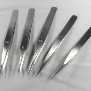 May include: Set of six stainless steel tweezers with different tip styles. The tweezers are labelled with numbers, including "00", "5", "4A", and "5".