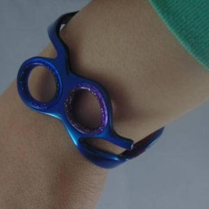 May include: A metallic blue cuff bracelet shaped like a pair of hair cutting scissors. The design features two circular finger holes with a shimmering purple interior finish, creating a distinct silhouette that wraps around the wrist area.